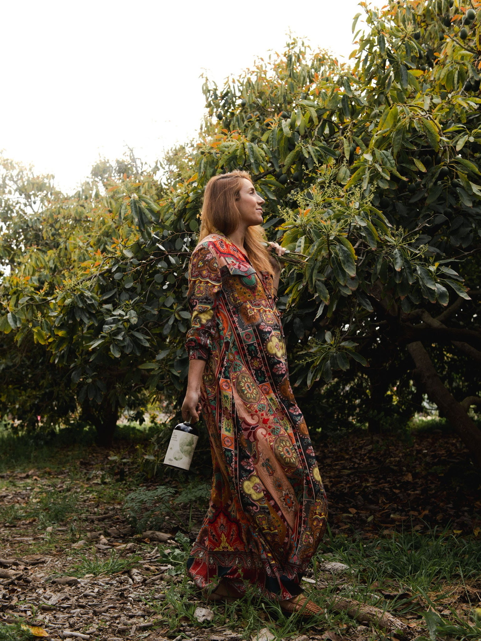 A woman in a long dress walking through an orchard, surrounded by blooming avocado trees.